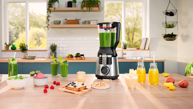 Uma bancada de cozinha de madeira com um liquidificador Bosch prateado e preto, rodeado por v&aacute;rios alimentos e bebidas saud&aacute;veis, como batidos verdes, frutas e tostas.