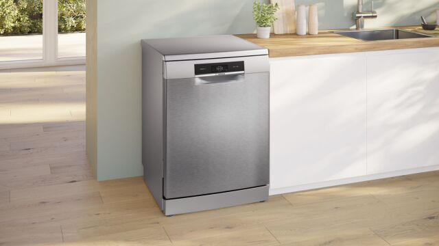 Uma m&aacute;quina de lavar loi&ccedil;a Bosch de cor cinza, com um painel de controlo preto, numa cozinha moderna com arm&aacute;rios brancos e bancada de madeira.