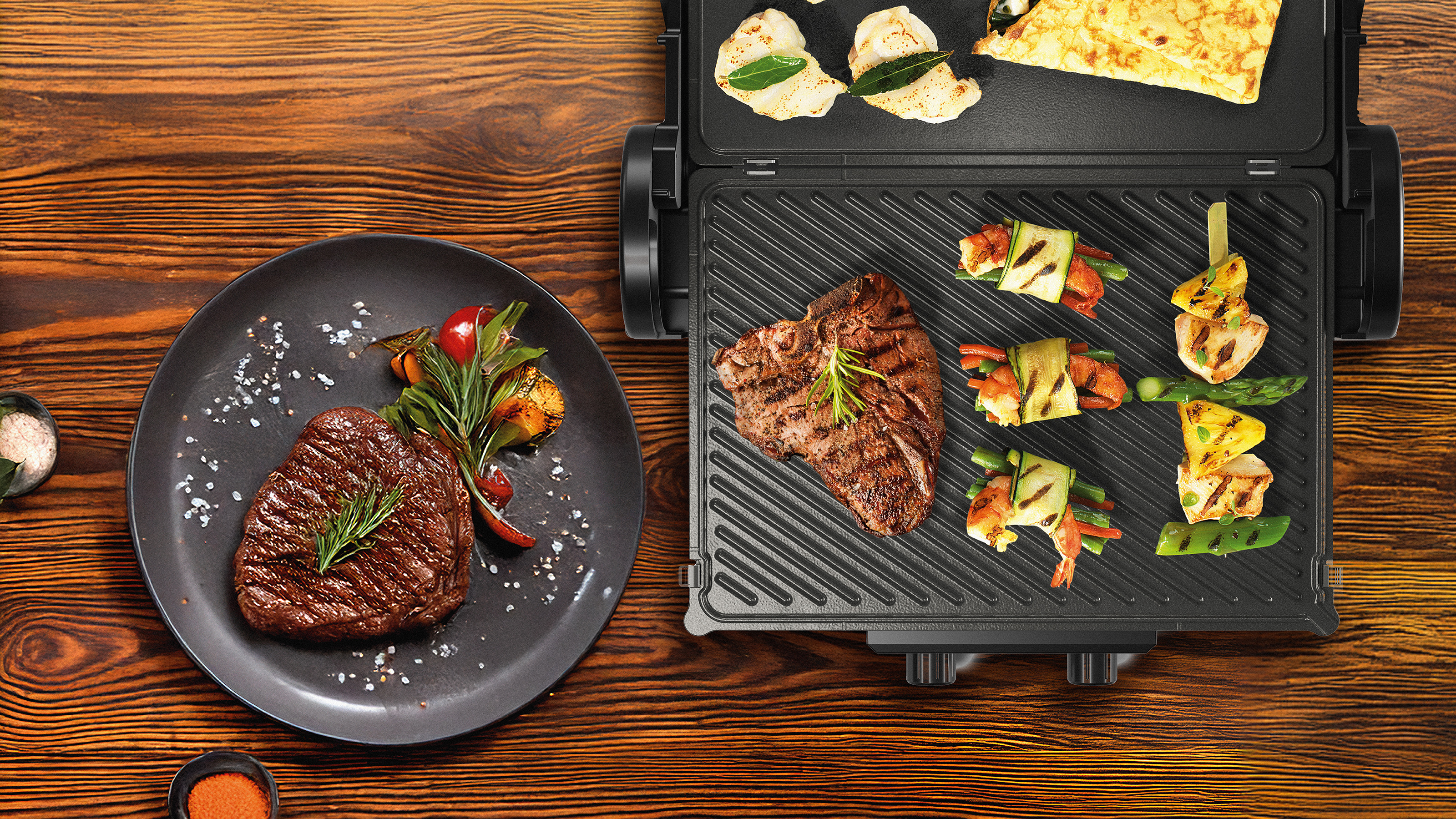 Um grelhador el&eacute;trico Bosch com bifes e espetadas de vegetais a grelhar, e um bife grelhado j&aacute; empratado com vegetais e alecrim.