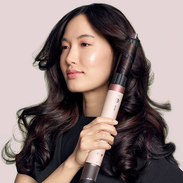 Una modelo morena usa el Airwrap Co-anda2x con accesorio moldeador para rizar su cabello, sobre un fondo rosa pálido.