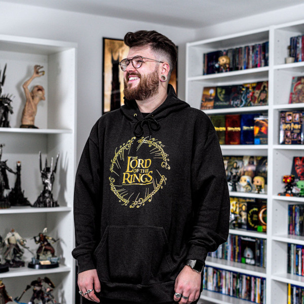 Lächelnder Mann mit schwarzem Hoodie, auf der Vorderseite das „The Lord of the Rings“-Logo in einem goldenen Kreis mit elbischer Schrift. Hoodie im Der-Herr-der-Ringe-Stil mit gemütlicher Passform.
