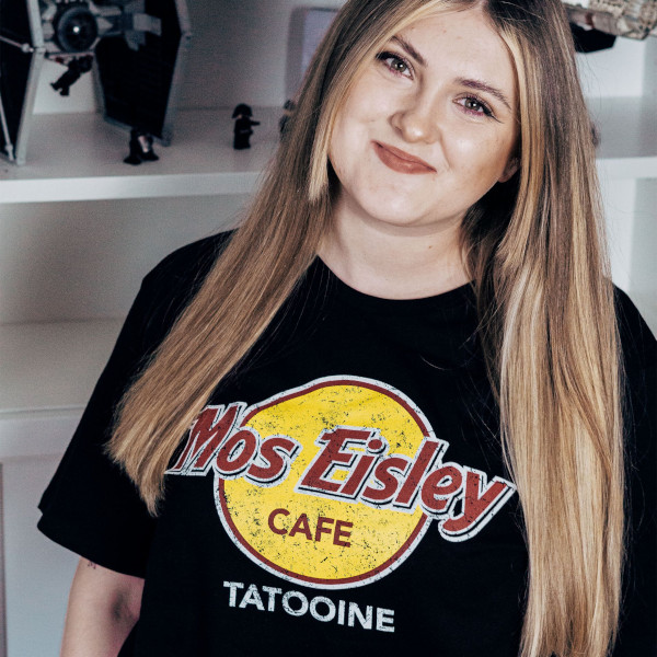 Person mit langen blonden Haaren trägt ein schwarzes T-Shirt mit rundem Logo und Schriftzug Mos Eisley Cafe Tatooine aus Star Wars. Das Motiv ist in Gelb, Rot und Weiß gehalten. Regal mit Star-Wars-Deko im Hintergrund.