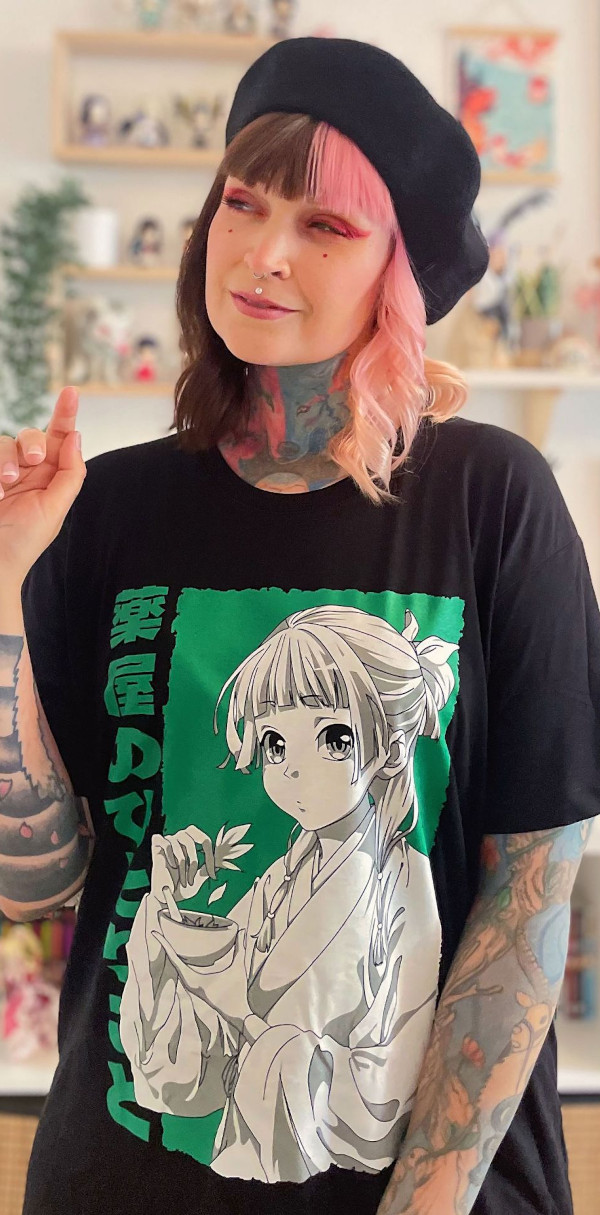 Person mit schwarz-rosa Haaren und schwarzer Baskenmütze trägt ein schwarzes T-Shirt mit großem Anime-Motiv. Auf dem Shirt ist eine gezeichnete Figur mit Schale in der Hand vor grünem Hintergrund zu sehen, japanische Schriftzeichen daneben. Innenraum mit Regal im Hintergrund.