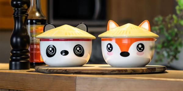 Zwei kleine Keramikschalen mit Deckel stehen auf einem Holzbrett in einer Küche. Die Schalen sind als niedliche Tiergesichter gestaltet, einmal Panda, einmal Fuchs, jeweils mit gelbem Hut.
