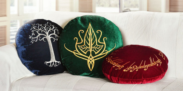 Drei runde Samtkissen liegen nebeneinander auf einem hellen Sofa. Die Kissen sind dunkelblau, grün und bordeauxrot und zeigen gestickte Symbole aus Der Herr der Ringe, darunter der Baum von Gondor, ein elbisches Zeichen und eine goldene Ringschrift.