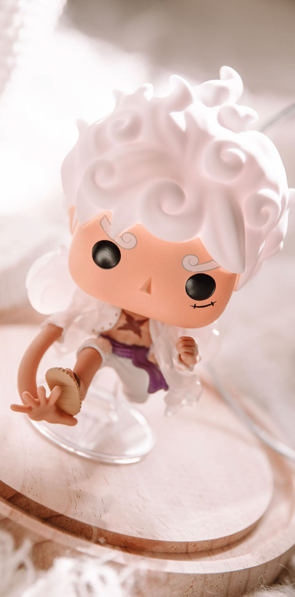 Stilisierte Vinyl-Figur mit großem Kopf, weißen Haaren und Flügeln schwebt leicht nach vorne gebeugt auf einer transparenten Halterung. Die Szene wirkt hell und wolkig.