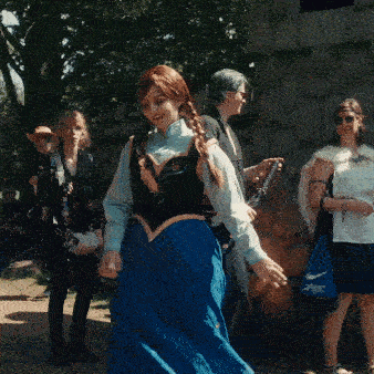 Person im Anna-Cosplay aus Disneys Frozen trägt ein blaues Kleid mit schwarzem Mieder und weißer Bluse und tanzt lachend im Freien. Im Hintergrund stehen weitere Festivalbesucher:innen, sommerliche Stimmung.
