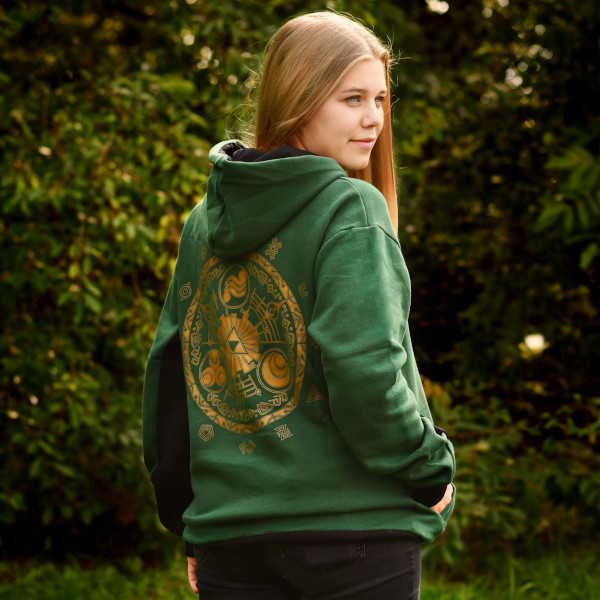 Person steht im Freien mit dem Rücken zur Kamera und trägt einen grünen Hoodie. Auf dem Rücken ist ein großes goldenes Symbol aus der The-Legend-of-Zelda-Welt zu sehen, inspiriert von Triforce und Ornamenten. Natürliche Umgebung mit Büschen im Hintergrund.