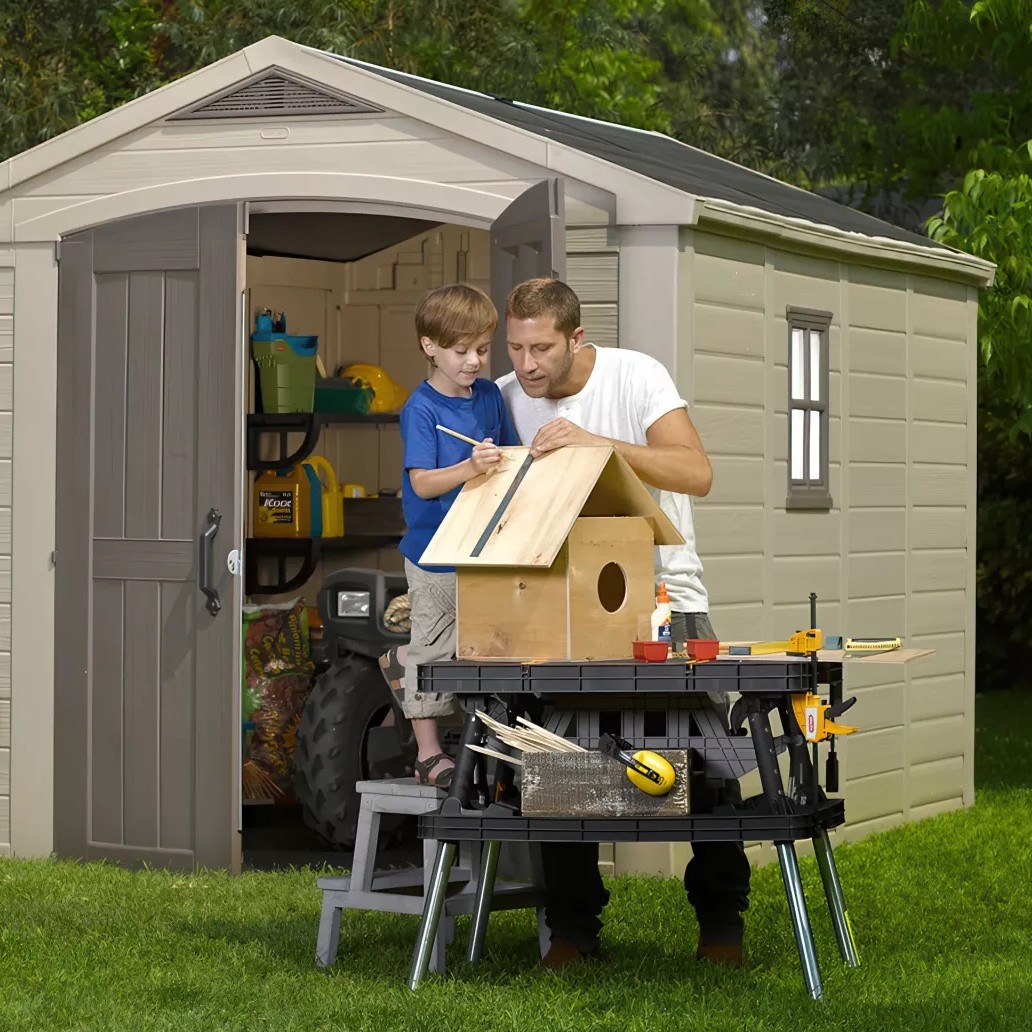 Man and boy building a wooden birdhouse on a workbench outside a garden shed, tools and grass visible