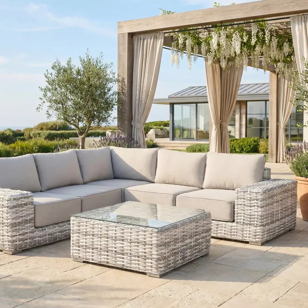 Light grey rattan outdoor corner sofa and glass-top coffee table under a pergola with curtains and hanging flowers, modern house and garden beyond