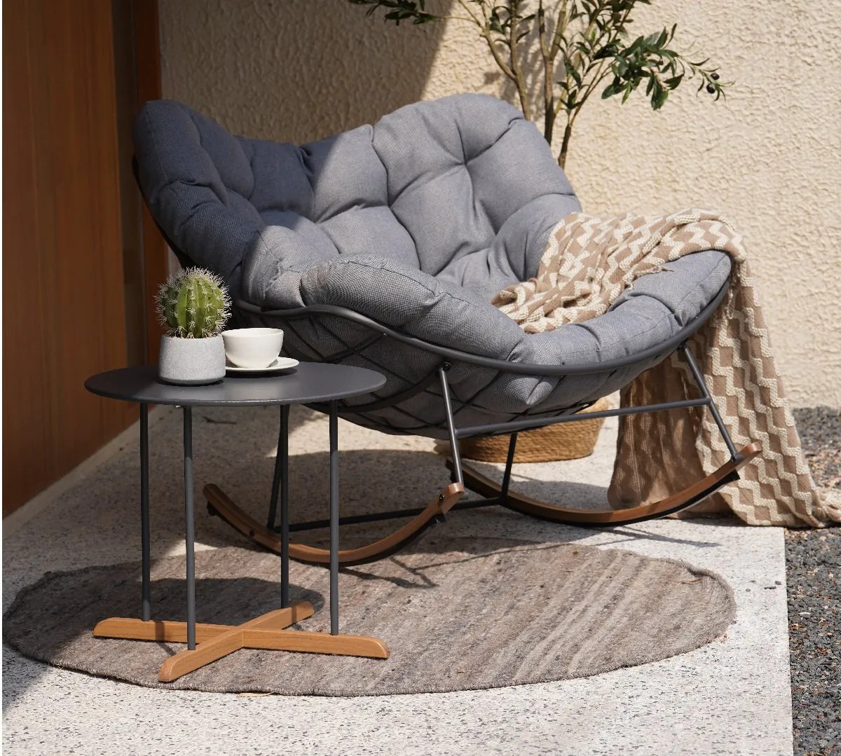 Cosy grey tufted rocking chair on sunlit patio with beige patterned throw, round rug and black side table holding a small cactus and coffee cup