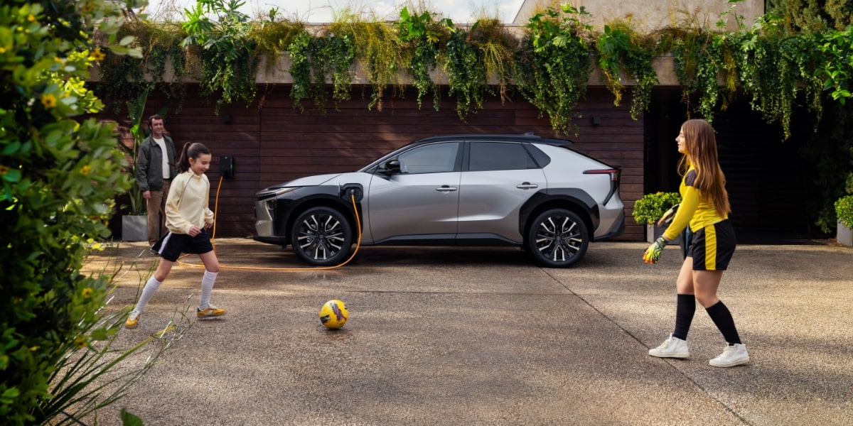 SUV Toyota bZ4X elétrico estacionado e duas crianças a jogar futebol à frente de uma casa moderna.