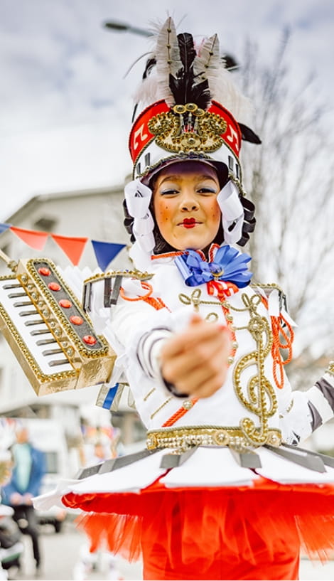 Pessoa fantasiada com traje festivo de banda/carnaval, em branco e vermelho, com chapéu alto decorado com penas,
maquilhagem colorida e um pequeno acordeão preso ao figurino, estendendo a mão em direção à câmara numa rua enfeitada para festa.