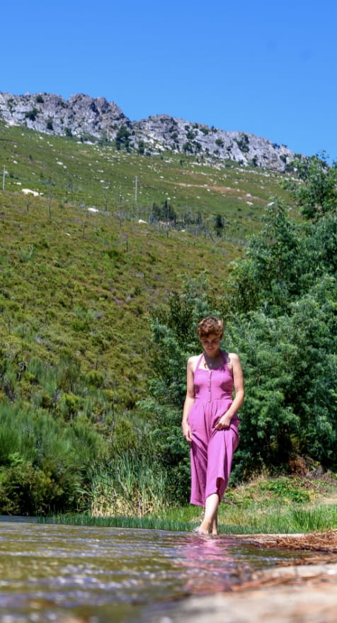 Rita Red Shoes vestida com um macacão rosa/lilás a caminhar descalça à beira de um rio, com uma encosta verdejante e montanhas rochosas ao fundo, sob um céu azul limpo.