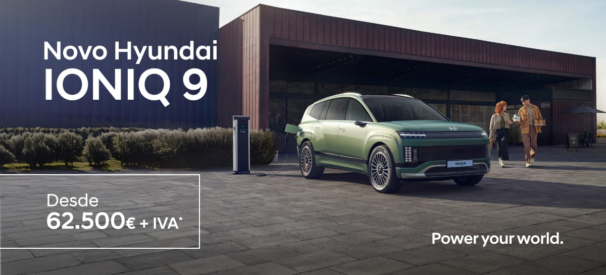 Hyundai IONIQ 9 verde estacionado junto a posto de carregamento, duas pessoas caminham perto de edifício moderno.