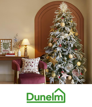 Dunelm: Festive room with a large decorated Christmas tree and a red chair with a patterned cushion. Nearby, a table holds a lamp and small decor items. Cozy and cheerful.