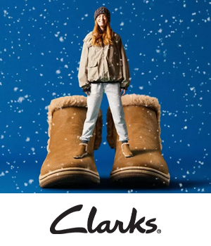 Clakrs - A smiling woman stands between oversized brown suede boots on a snowy blue background. She's wearing a cozy winter outfit.