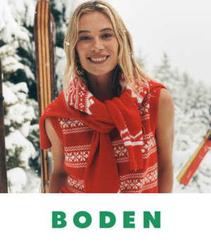 Boden - Smiling woman in a red and white winter sweater stands amidst snowy trees and wooden skis, conveying a cozy, cheerful winter vibe.