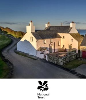 National Trust Holidays: Aerial view of a serene countryside setting with a large white house featuring two chimneys, winding road, stone walls, and a distant sea under clear blue skies.