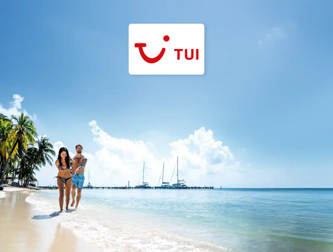 TUI - A couple in swimwear walks along a sandy beach near palm trees and sailboats in the ocean