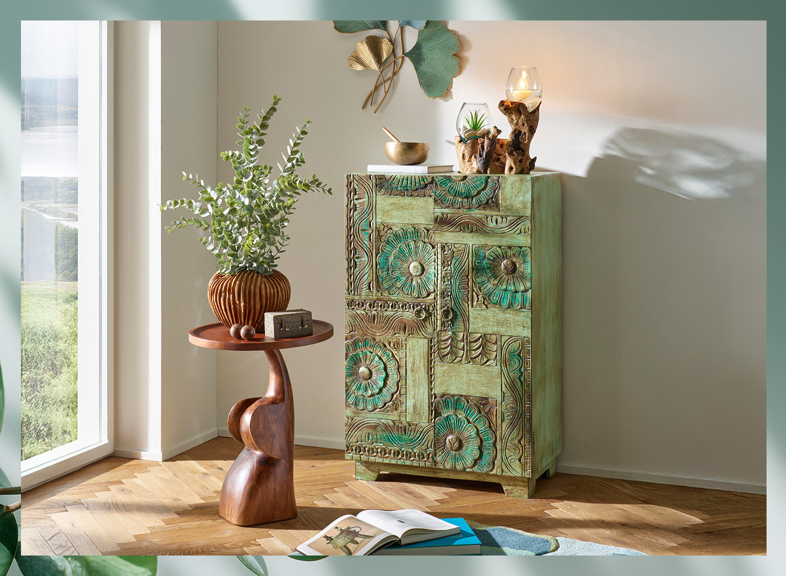  Ein grüner, kunstvoll verzierter Schrank steht neben einem kleinen, braunen Beistelltisch mit einer Vase und Pflanzen, im hellen Raum mit Fenster.