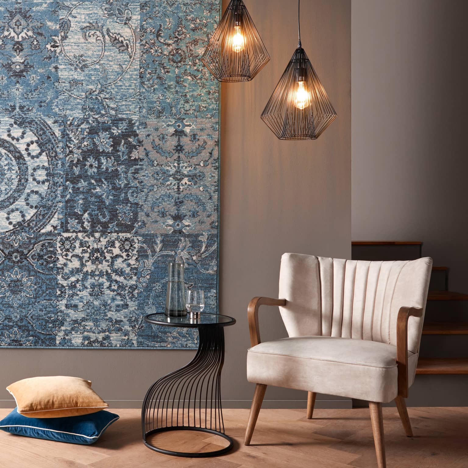 Gemütliche Sitzecke mit beigem Sessel, kleinem schwarzen Tisch, zwei Kissen auf dem Boden und blau gemustertem Wandteppich.