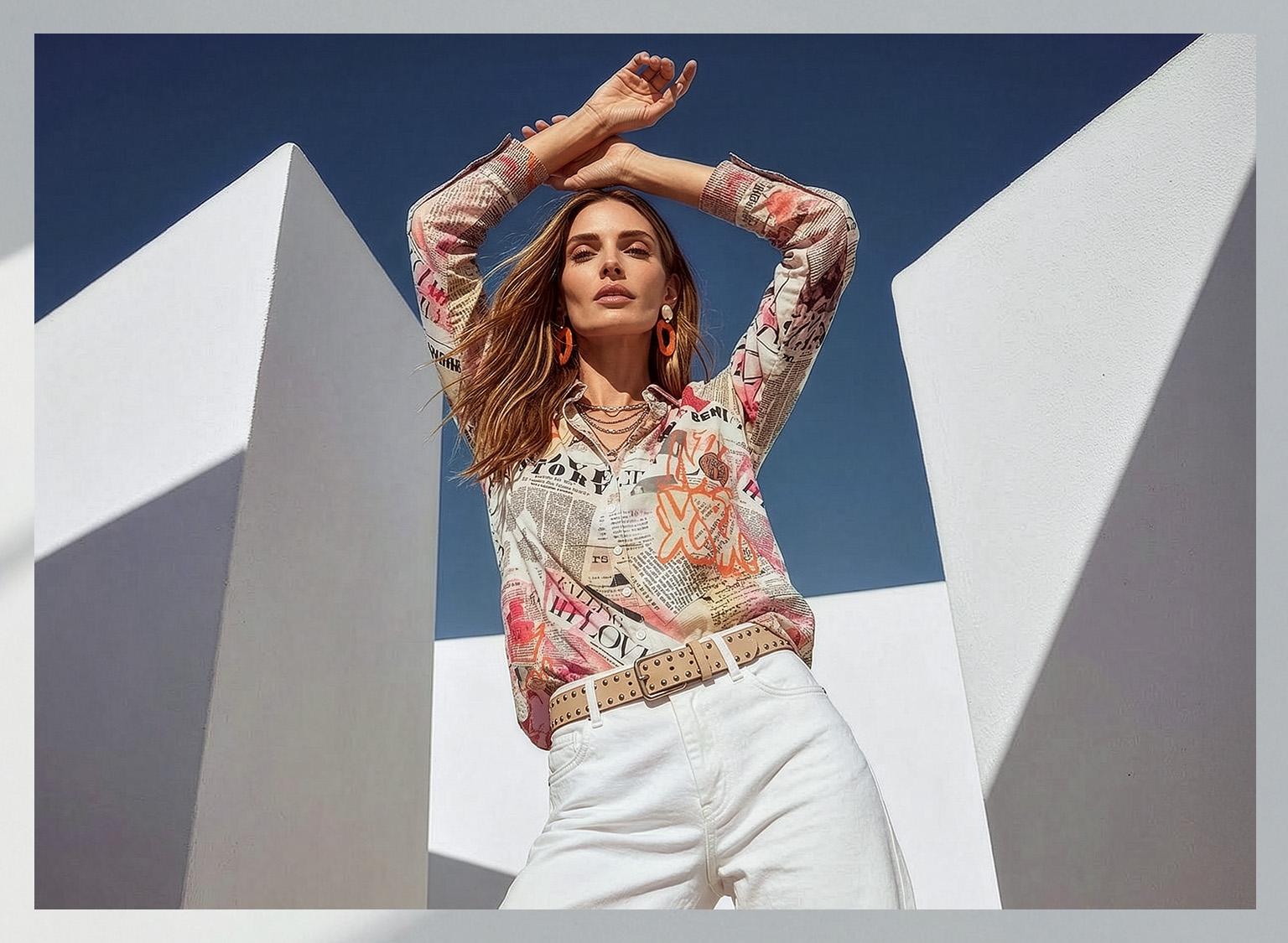 Ein Model steht vor weißem, modernem Gebäude, trägt eine bunte Bluse mit Schriftmustern und weiße Hosen, die Arme sind über dem Kopf verschränkt.