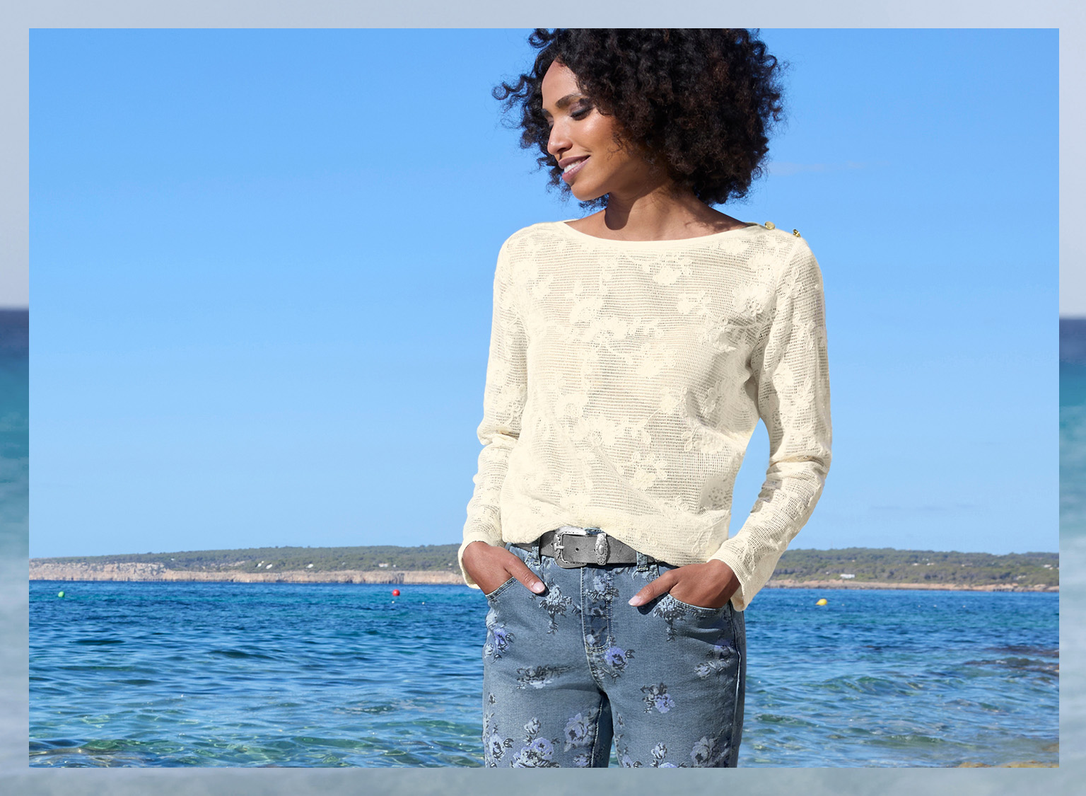 Ein Model steht am Meer und trägt einen hellen, strukturierten Pullover und Jeans mit floralem Muster. 
