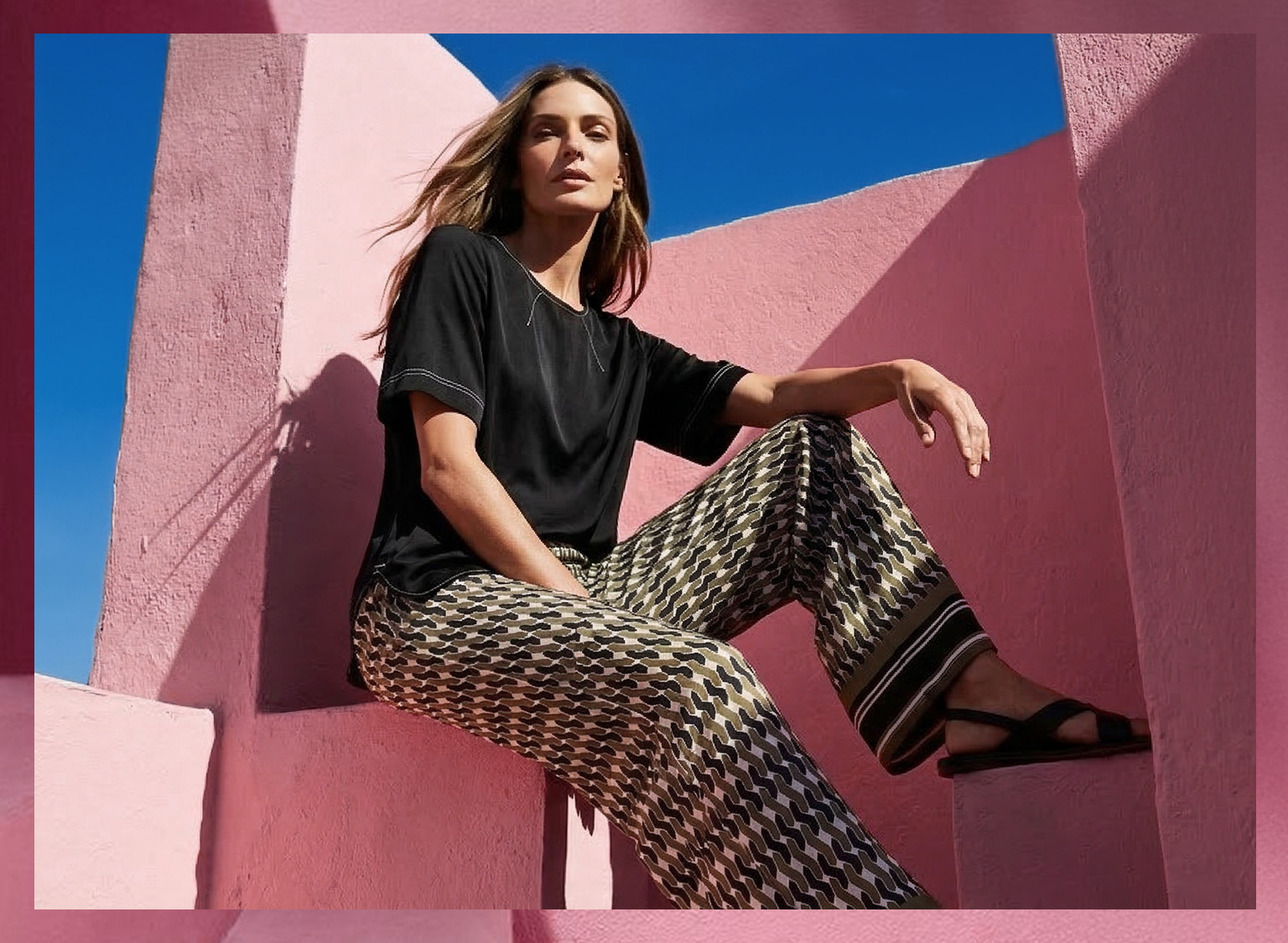 Ein Model sitzt auf einer rosa Treppe und trägt ein schwarzes T-Shirt mit kurzen Ärmeln und gemusterten, weit geschnittenen Hosen in Schwarz, Weiß und Beige.