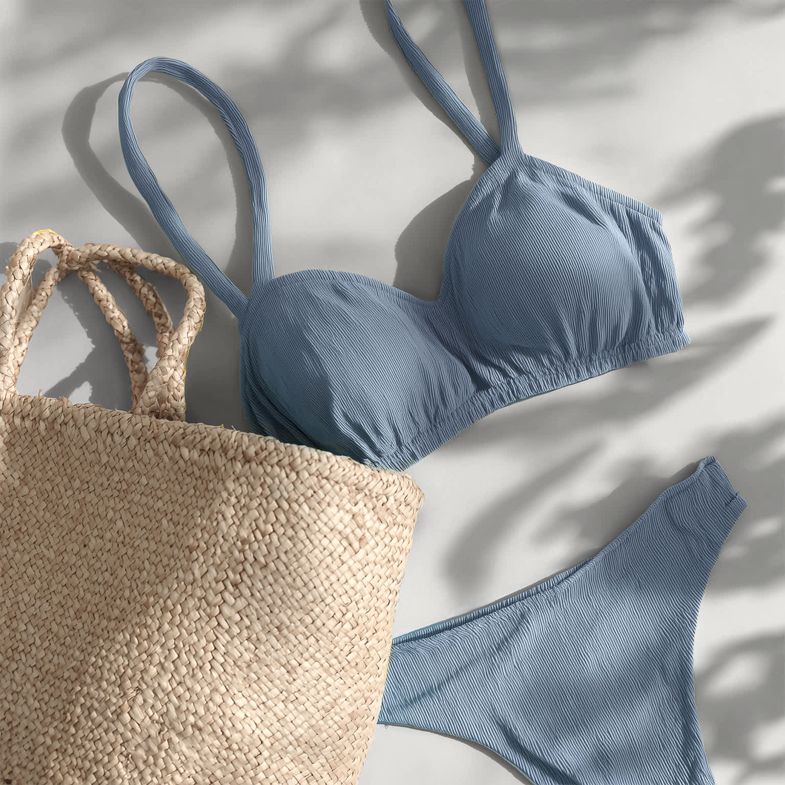 Hellblauer Bikini mit Textur liegt neben einem geflochtenen Strohtasche auf hellem Untergrund mit Schatten.