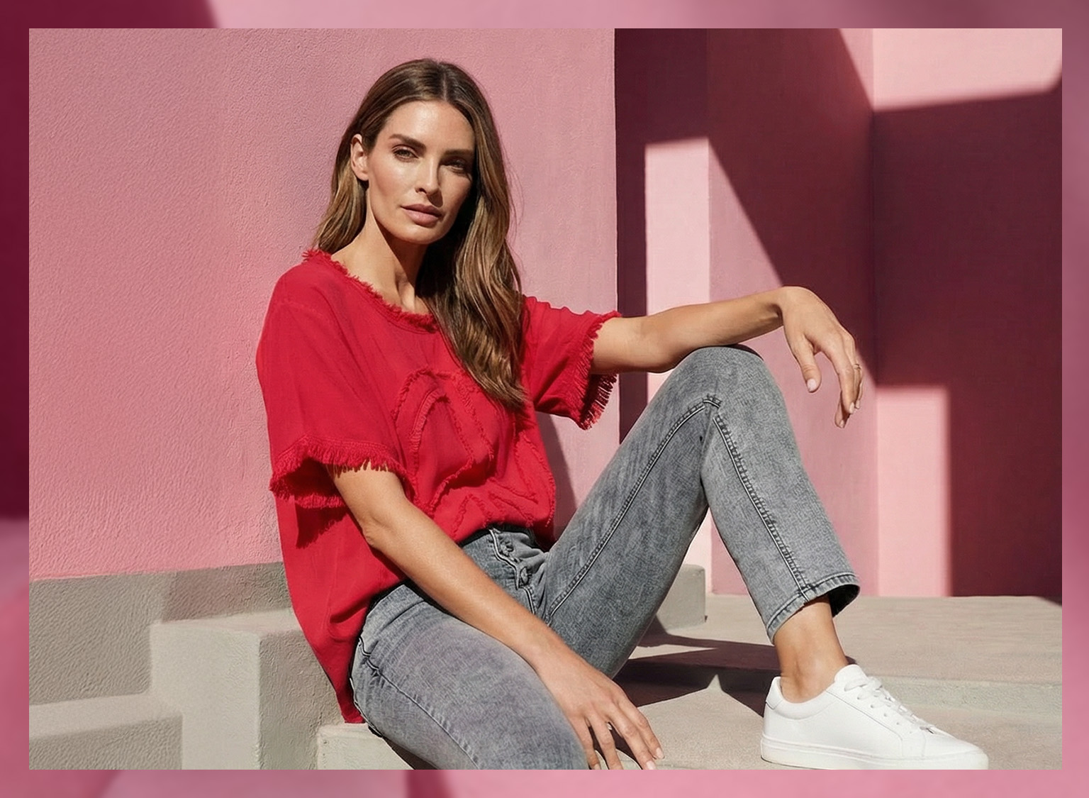 Ein Model sitzt auf einer Treppe vor einer pinkfarbenen Wand, trägt ein rotes, kurzärmeliges Shirt, graue Jeans und weiße Sneaker.