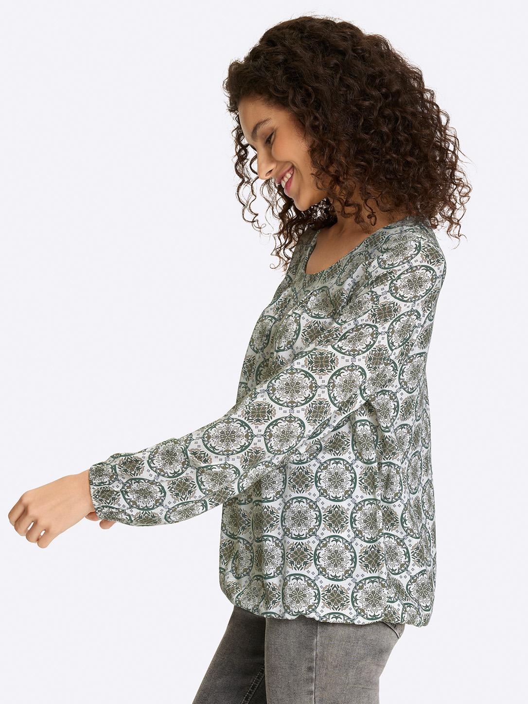 Ein Model trägt eine langärmelige Bluse mit grün-weißem, rundem Muster, dazu graue Hosen, vor weißem Hintergrund. 
