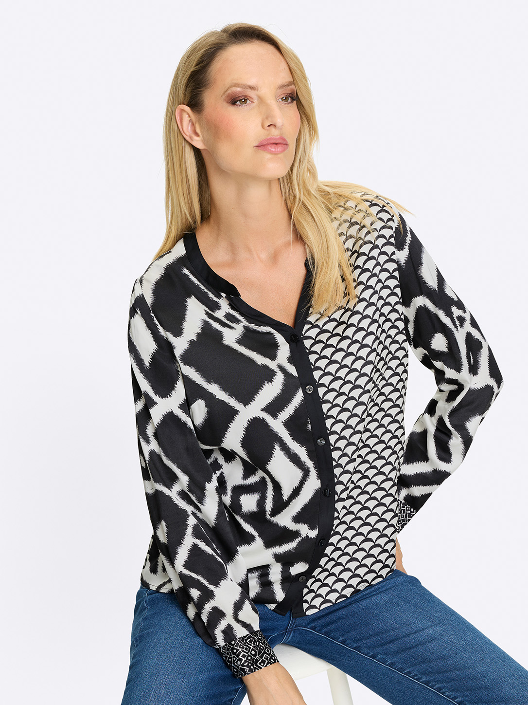 Ein Model trägt eine langärmelige Bluse mit schwarz-weißem, gemischtem Muster, dazu blaue Jeans, vor weißem Hintergrund. 