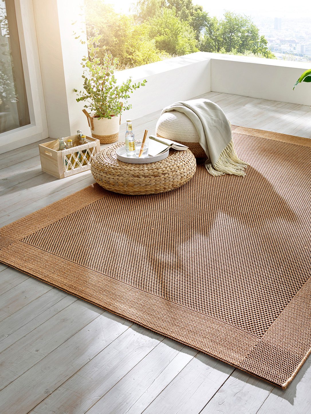 Heller Balkon mit großem gewebtem Teppich, runder Korbtisch, Pouf mit Decke, Pflanze und Getränken in der Sonne.