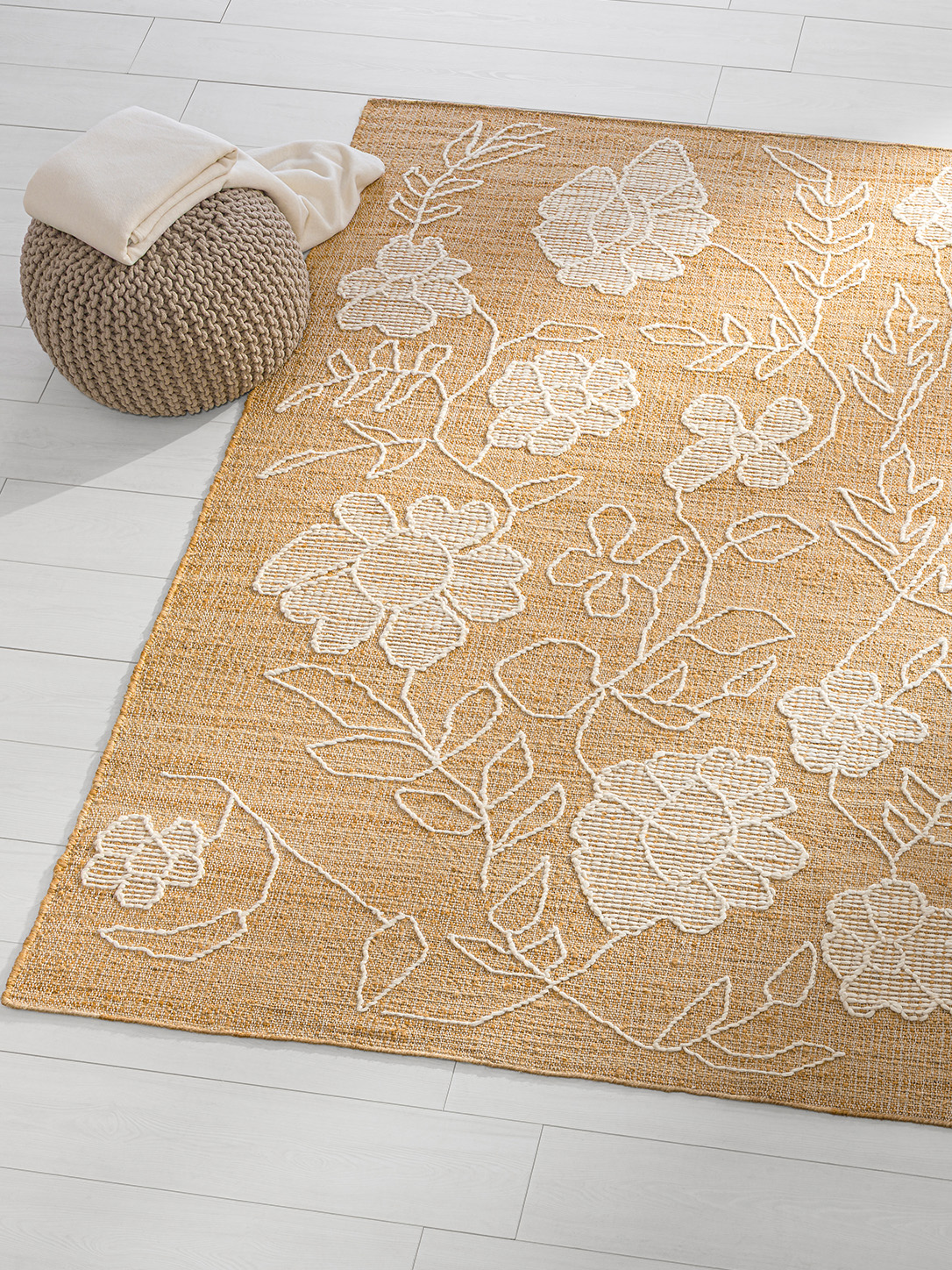 Heller Teppich mit weißem Blumenmuster, daneben ein grauer Strick-Pouf mit einer weißen Decke darauf.