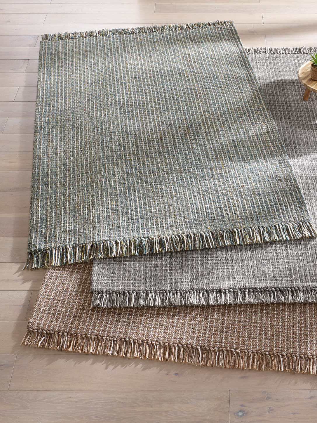 Drei übereinanderliegende Teppiche mit Fransen, in verschiedenen gedeckten Farben und feinem Streifenmuster auf hellem Holzboden. 