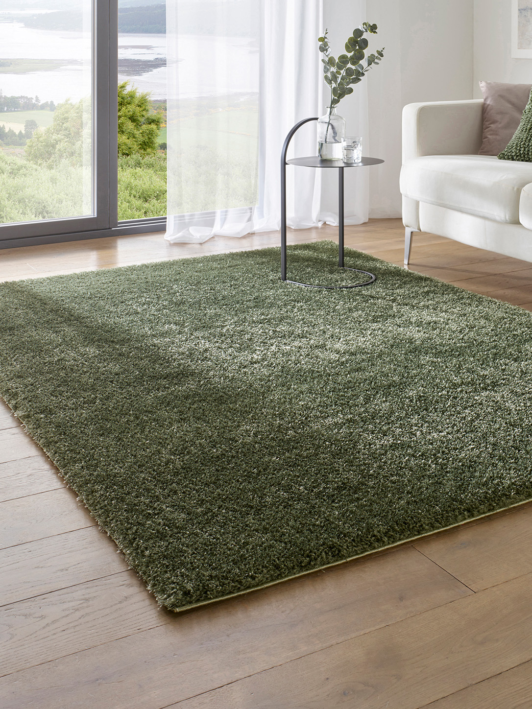  Grüner, dichter Teppich auf hellem Holzboden, daneben ein weißes Sofa und ein kleiner runder Beistelltisch mit Vase.