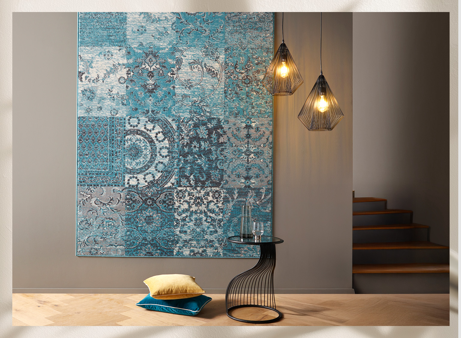 Moderner Raum mit blau-grau gemustertem Wandteppich, zwei hängenden Drahtlampen, kleinem runden Tisch und zwei Kissen auf hellem Holzboden.