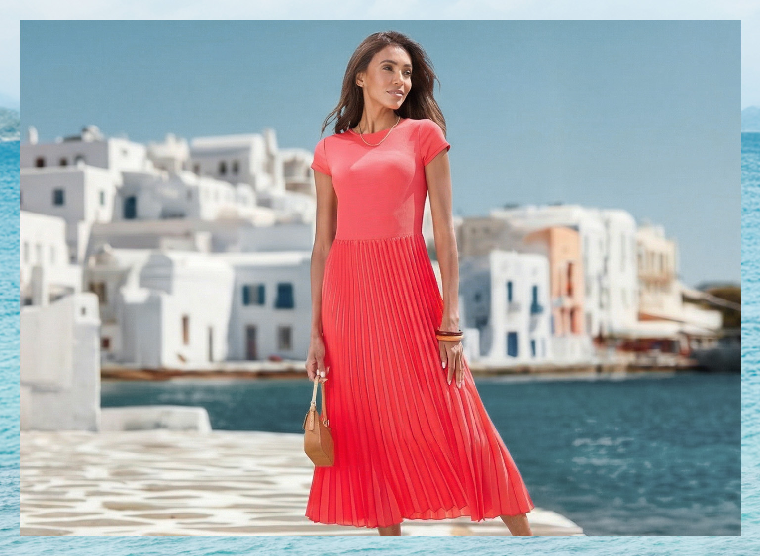 Ein Model steht am Wasser vor weißen Häusern, trägt ein rotes, plissiertes Maxikleid und hält eine kleine Tasche.