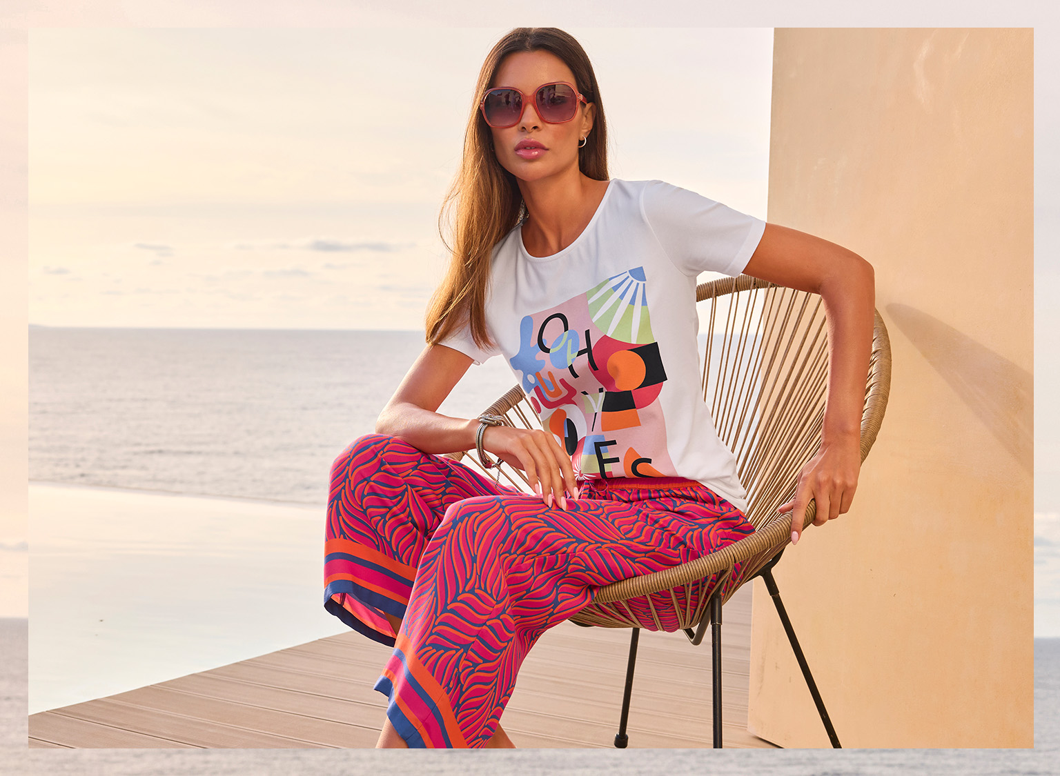 Ein Model sitzt auf einem Stuhl am Wasser, trägt ein weißes T-Shirt mit buntem Print und weite pink-rote Hose mit Muster. 