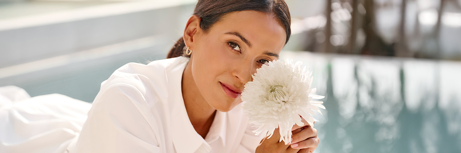 Ein Model mit zusammengebundenen Haaren hält eine weiße Blume in der Hand und trägt ein weißes Oberteil. Im Hintergrund Wasser.