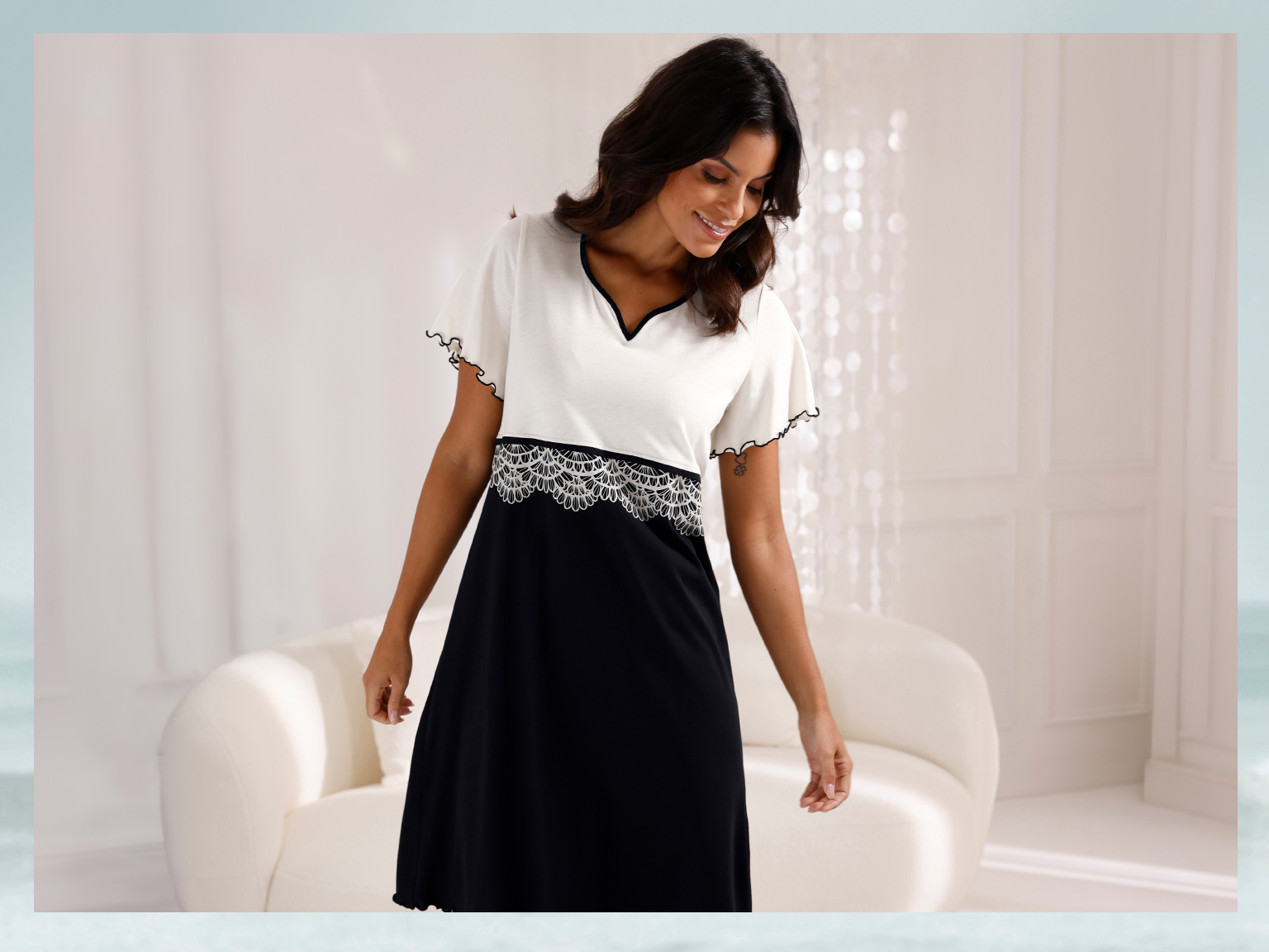 Ein Model trägt ein schwarz-weißes Kleid mit kurzen Ärmeln und einer Spitzenborte an der Taille. Im Hintergrund ein heller Raum.