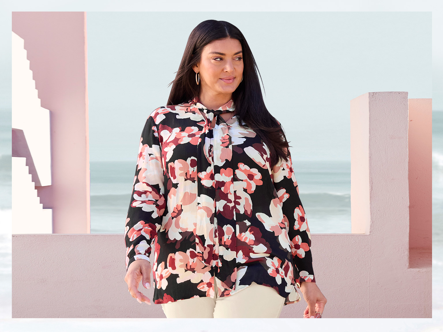 Ein Model trägt eine langärmelige Bluse mit großem, buntem Blumenmuster und helle Hose. Im Hintergrund Architektur in Rosa und das Meer.