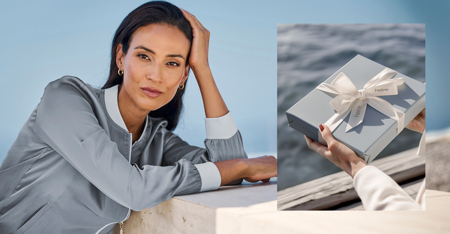Ein Model lehnt entspannt an einer Mauer, daneben eine Hand, die eine graue Geschenkbox mit weißer Schleife hält.