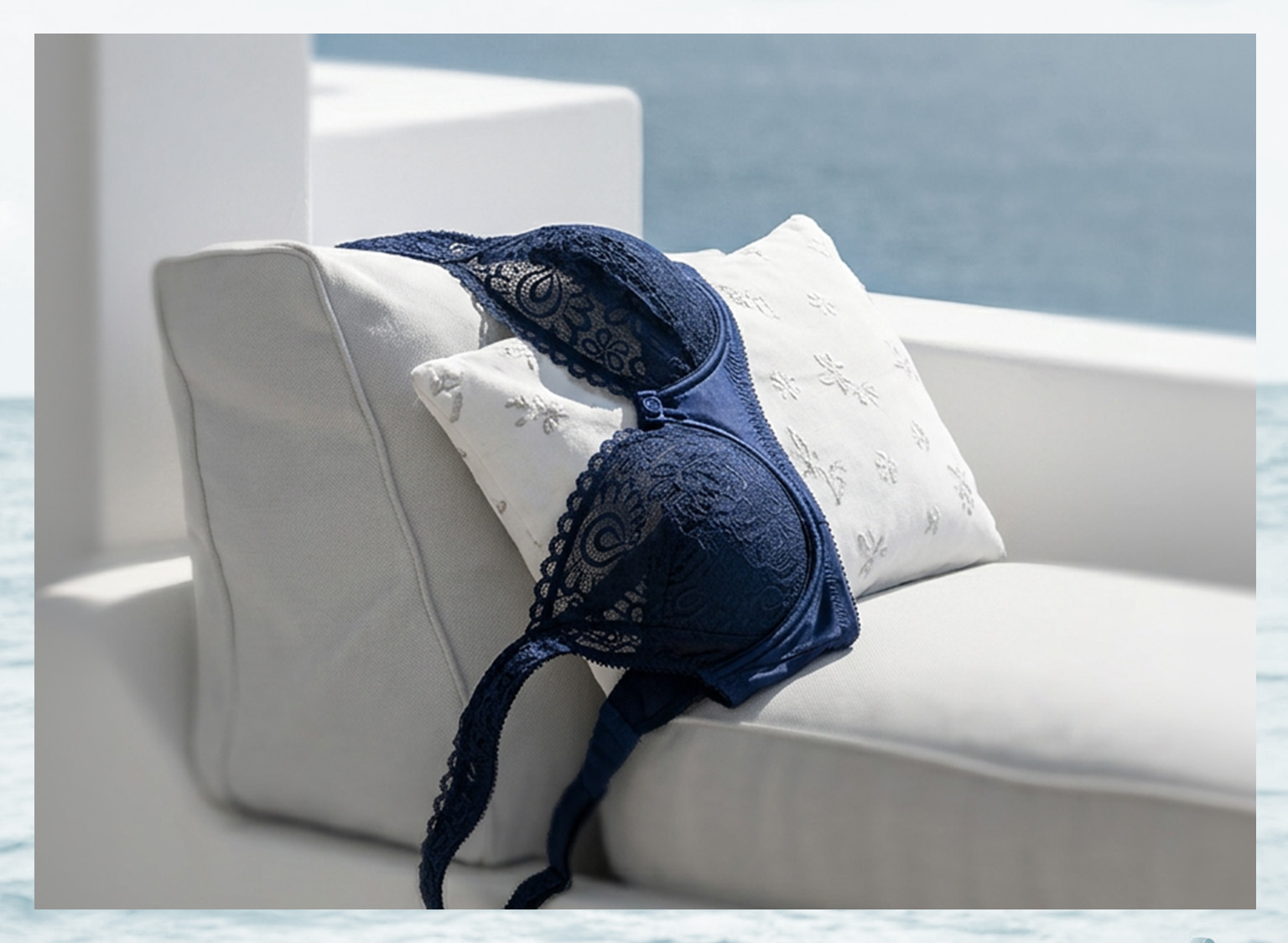 Ein dunkelblauer Spitzen-BH liegt auf einem weißen Kissen auf einem hellen Sofa, im Hintergrund Meer und Himmel.