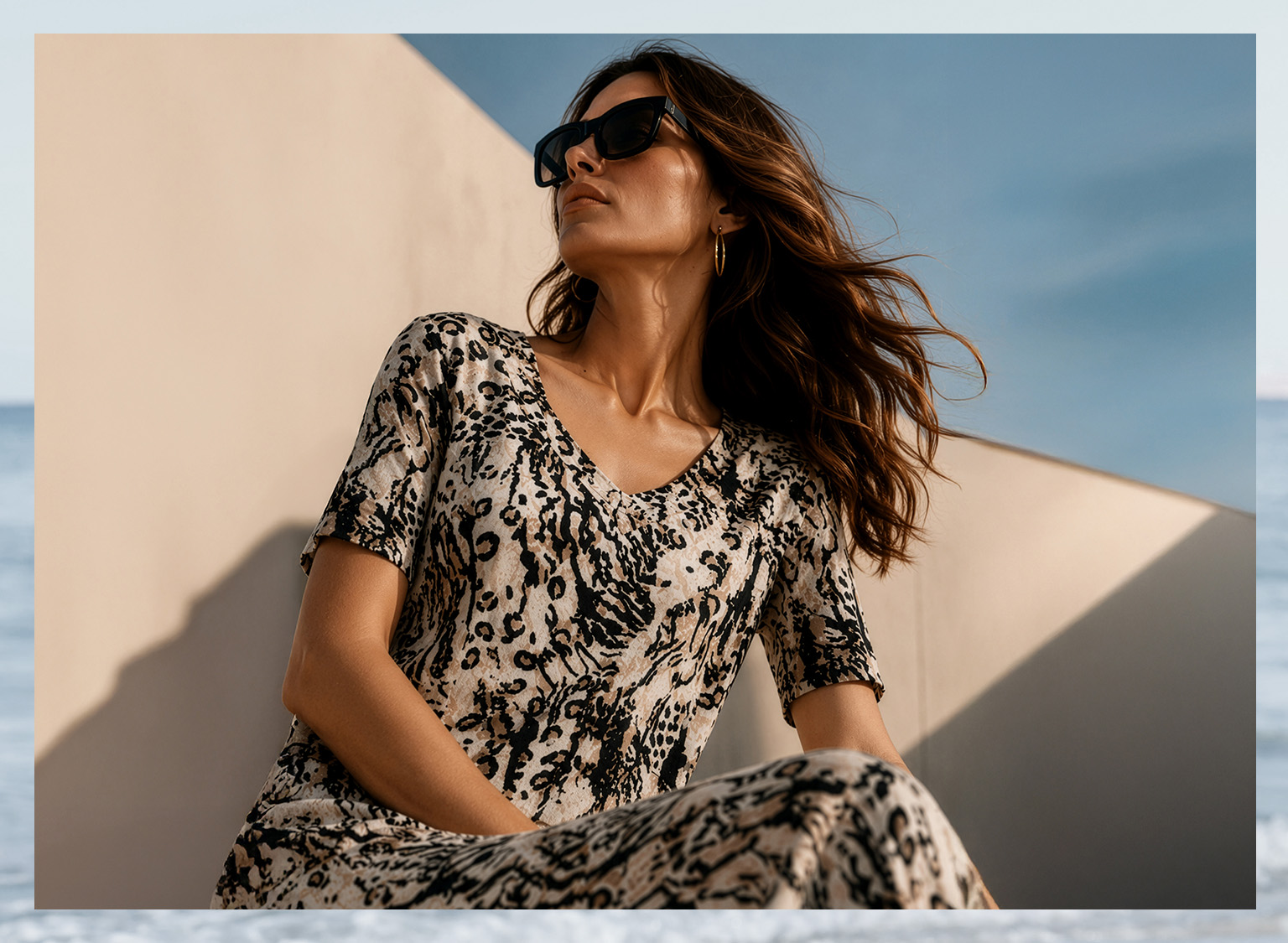 Ein Model sitzt draußen, trägt ein kurzärmeliges Kleid mit Leopardenmuster und Sonnenbrille. Der Himmel ist blau, im Hintergrund eine helle Wand.