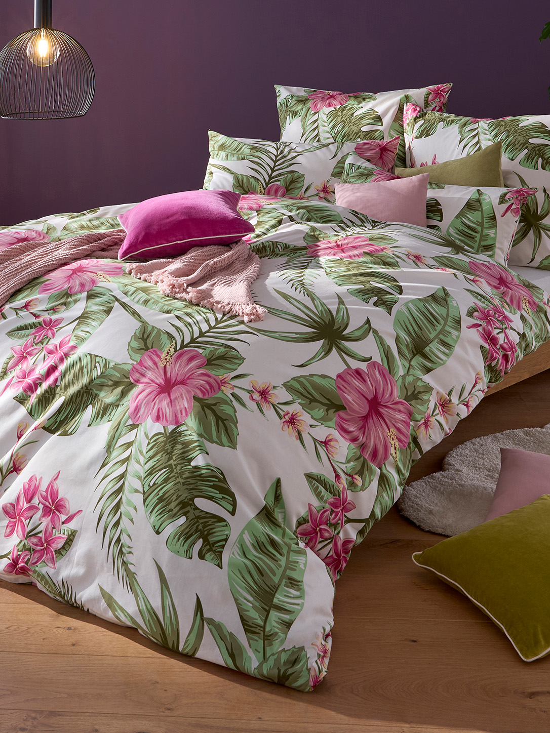 Ein Bett ist mit Bettwäsche in Weiß mit großem grünem Blatt- und pinkem Blumenmuster bezogen. Verschiedene Kissen in Rosa und Grün liegen darauf. Im Hintergrund ist eine violette Wand.
