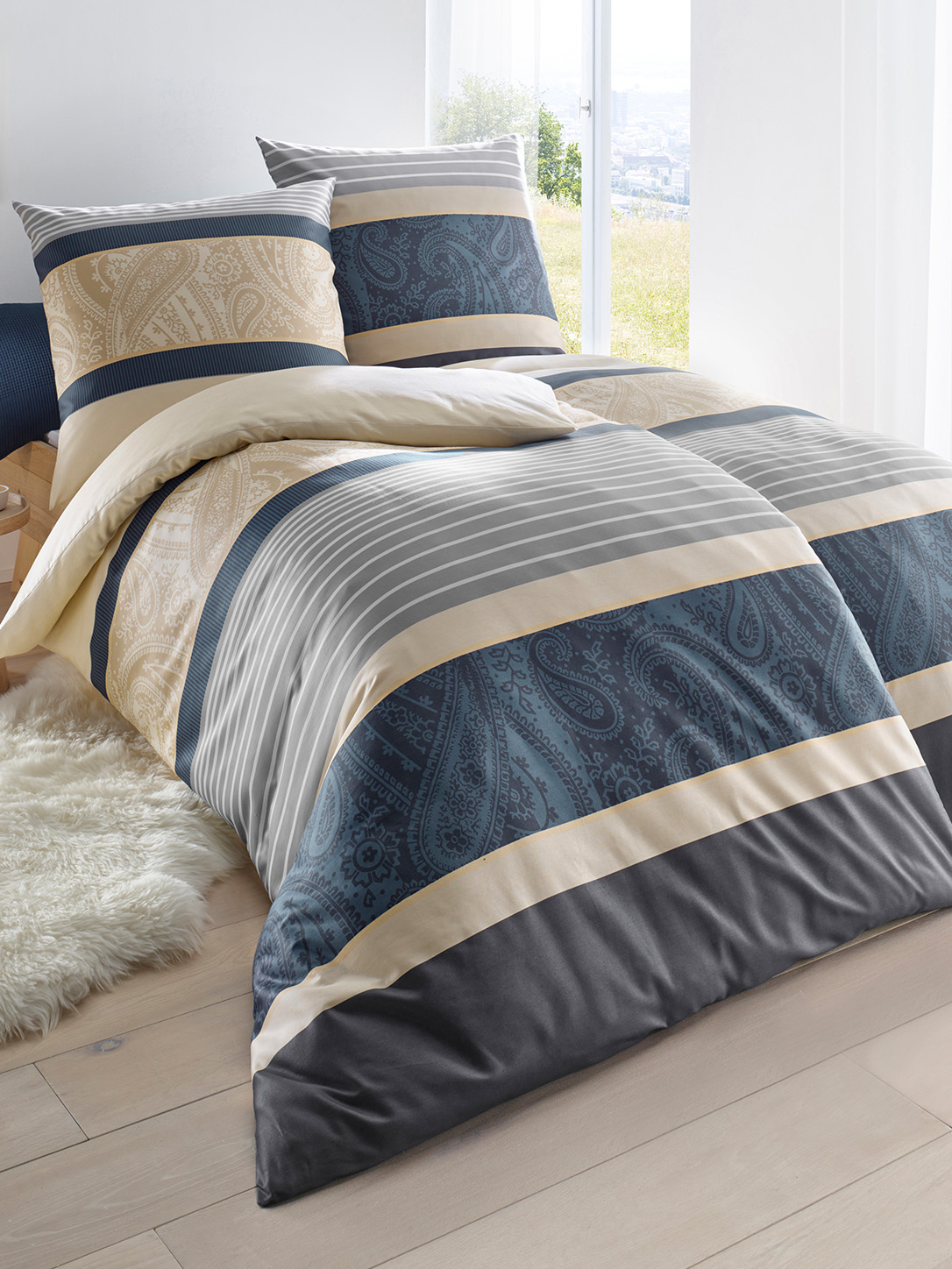  Ein Bett ist mit Bettwäsche in Streifenmuster bezogen, die Farben sind Blau, Beige, Grau und Weiß mit Paisley-Details. Das Zimmer ist hell mit einem Fenster und einem weißen Teppich.