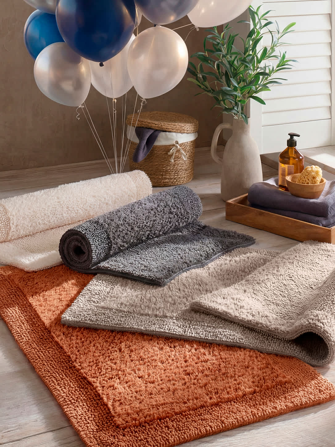  Mehrere flauschige Badteppiche in Beige, Grau und Orange liegen übereinander auf einem Holzboden, daneben ein Tablett mit Handtüchern, Seifenspender und Schwamm, im Hintergrund Ballons und ein Korb.