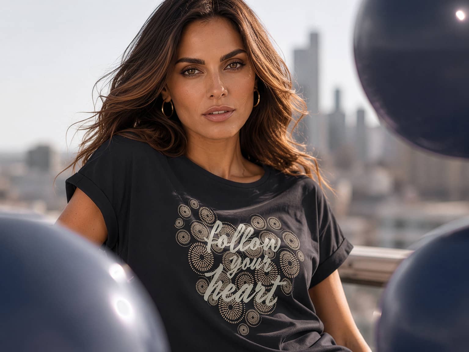 Ein Model trägt ein schwarzes T-Shirt mit goldfarbenem Herz und Schriftzug 'follow your heart', im Hintergrund Stadt und große schwarze Luftballons.